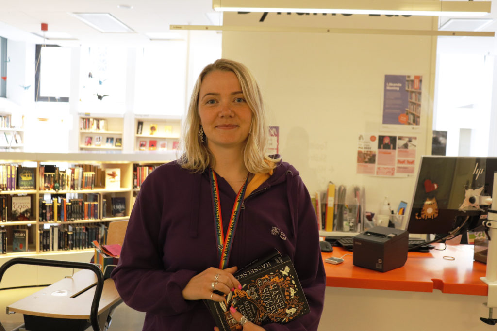 Alma Kilander är bibliotekarie på Göteborgs stadsbibliotek. Hon står med en bok i handen och ler. Bakom henne finns hyllor med böcker. 