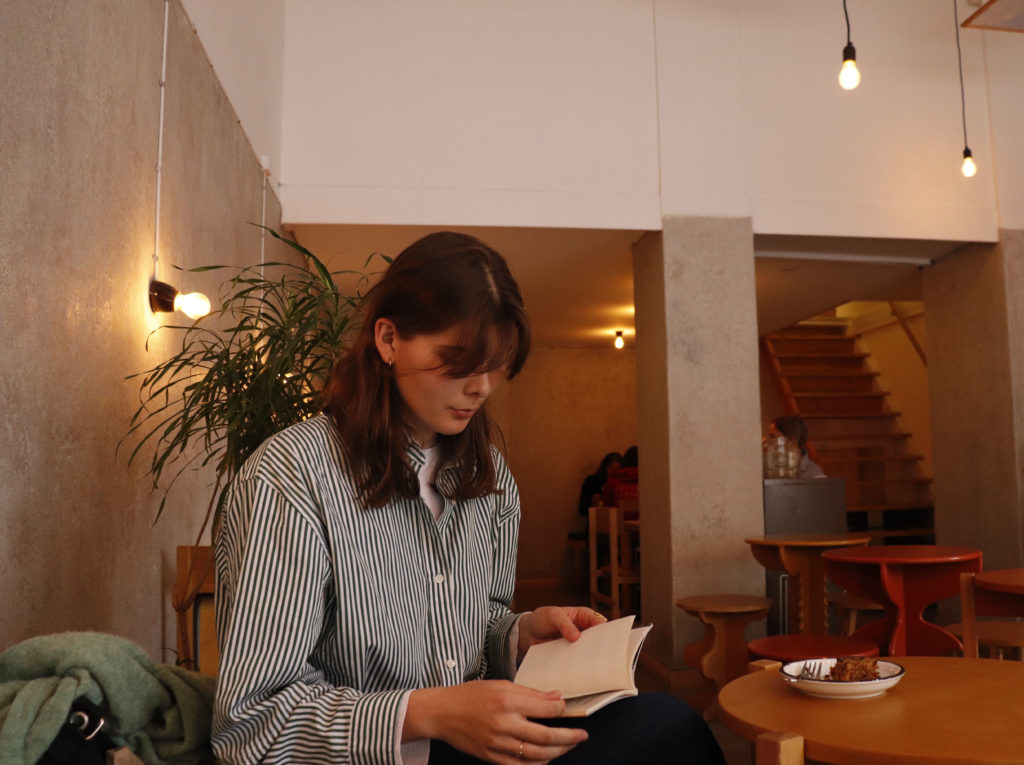 Julia sitter på ett café i Göteborg och läser ur en bok. I taket hänger svaga lampor och rummet är fullt av trästolar. 