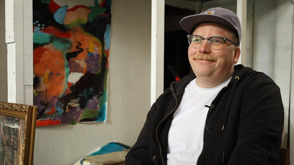 Bild på graffitimålaren och konstnären Code26 i hans ateljé. Erik är blond och har en blond mustasch. Han har på sig en grå keps, glasögon och en vit T-shirt och en svart hoodie. I bakgrunden syns en abstrakt målning och en guldfärgad tavelram.