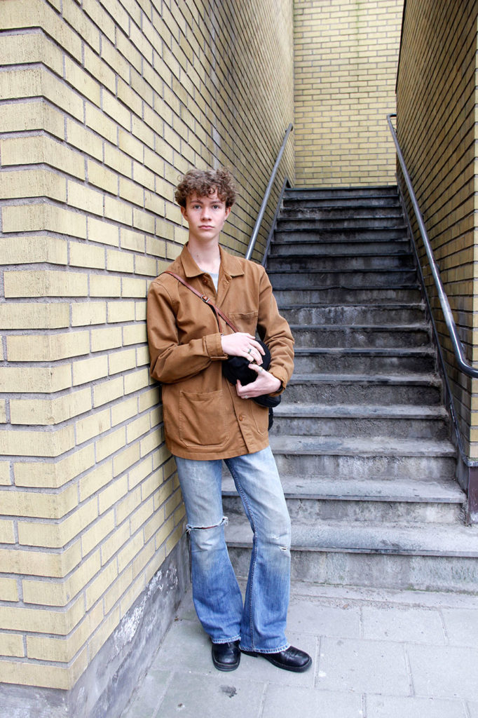 Olle står mot en vägg och kollar in i kameran. Han är klädd i second hand-kläder från topp till tå, bortsett från den grå tröjan. Han har på sig ett par blå utsvängda jeans, en brun/orange jacka och svarta skor. I händerna håller han sina nyfunna svarta skinnloafers.