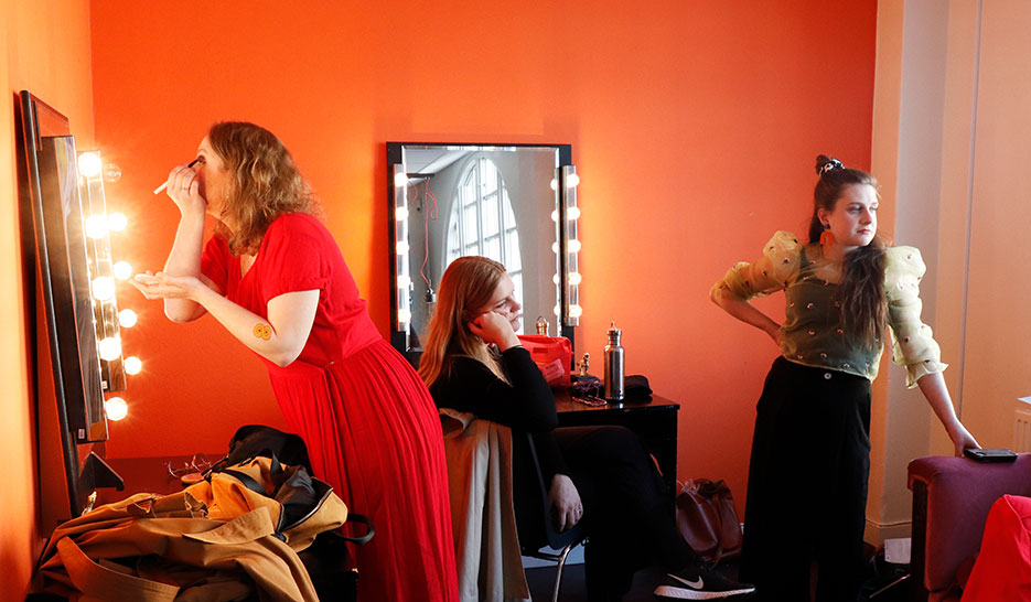 Amanda står framåtlutad över ett sminkbord mot en spegel. Hon har en borste i handen och lägger på ögonskugga. Bakom henne sitter Sara i en stol, och tittar på något utanför bild. Elin står bredvid och lutar sig mot soffan.