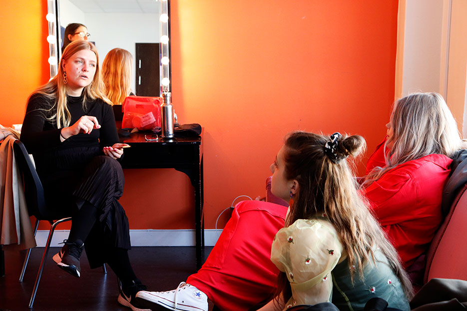 Gabriella och Elin sitter i en soffa och tittar snett upp på Sara, som sitter i en stol mittemot soffan. Hon har telefonen i handen, och sjunger.