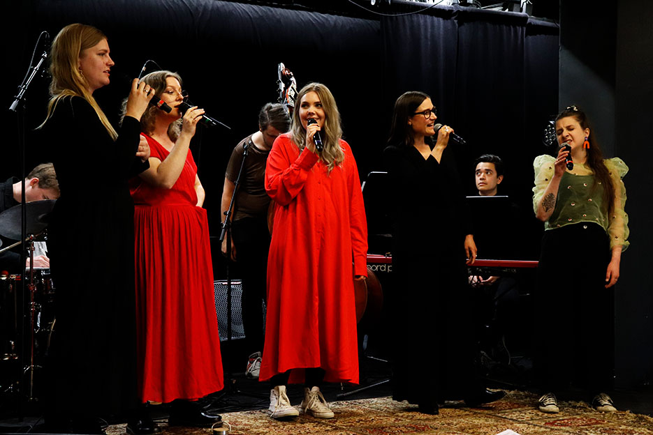 Fem kvinnor står på scen och sjunger i mikrofoner.