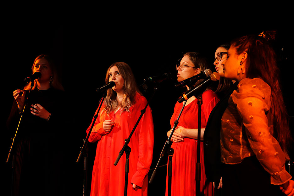 Fem kvinnor sjunger i mikrofoner med stativ.